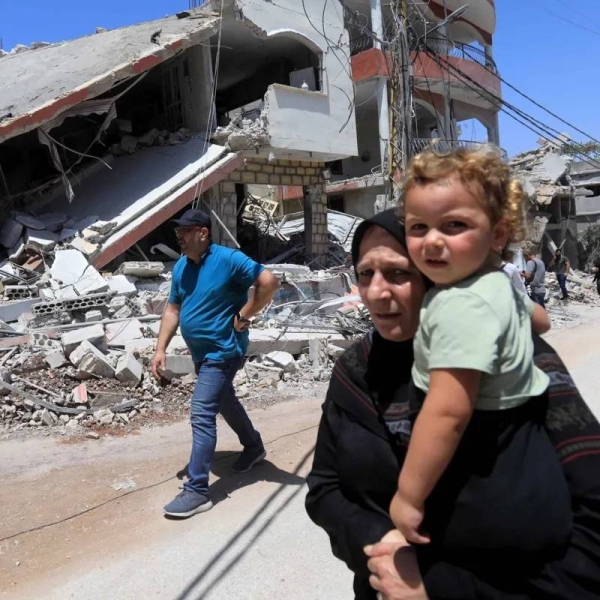 A woman carries a small girl while walking down a street in front of a destroyed building