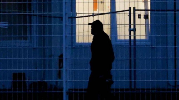 A man in a baseball cap walks past a window at night. He's silhouetted against the building and the soft white light coming from the window