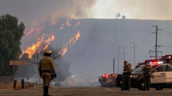 Emergency crews patrol along a street as flames and smoke billows into the air in the distance