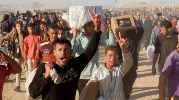 A Palestinian man and boy in the foreground look upset and make the 'peace' sign while carrying aid, including a box branded GHF for the Gaza Humanitarian Foundation