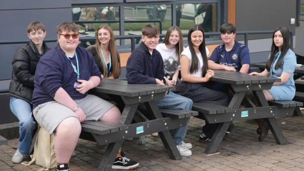 A group of male and female students, all aged 17, sit together, smiling, at two picnic tables in front of their college building