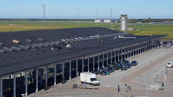 An airport terminal with a black roof is pictured from above, with a series of black vehicles parked in front of the building. The airport control tower can be seen in the distance