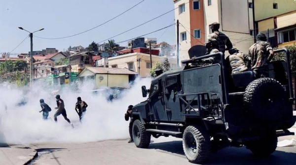 Riot police in a heavily armoured bear-cat kind of vehicle drive behind three protesters who are running away through tear gas smoke